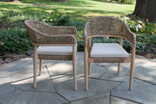 Load image into Gallery viewer, Two wicker chairs with white cushions on a stone patio with greenery in the background