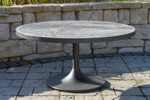 Load image into Gallery viewer, Round outdoor table with a stone-like finish on a patio with a stone wall background