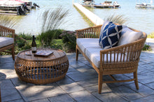 Load image into Gallery viewer, Outdoor patio setting with wicker furniture and a bottle of wine by a waterfront.
