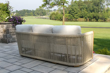 Load image into Gallery viewer, Outdoor wicker sofa with cushions on a patio area with greenery in the background
