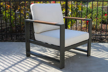 Load image into Gallery viewer, Chair with white cushions on a patio area with a black metal fence and greenery in the background.
