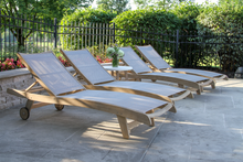 Load image into Gallery viewer, Four wooden lounge chairs on a stone patio with greenery in the background
