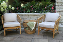 Load image into Gallery viewer, Two wicker chairs with white cushions and a woven table in an outdoor setting with flowers and a stone wall.
