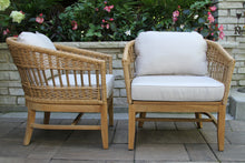 Load image into Gallery viewer, Two wicker chairs with white cushions on a patio with flowers and a stone wall in the background.
