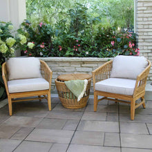 Load image into Gallery viewer, Two wicker chairs with white cushions and a matching table on a patio with greenery in the background.
