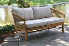 Load image into Gallery viewer, Wicker outdoor sofa with beige cushions on a stone patio by a body of water.
