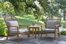 Load image into Gallery viewer, Two cable wicker and wood chairs with cushions and a small table on a patio with greenery in the background.
