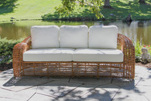 Load image into Gallery viewer, Wicker outdoor sofa with white cushions in a garden setting.

