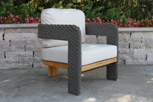 Load image into Gallery viewer, Outdoor chair with woven armrests and white cushions on a stone patio.
